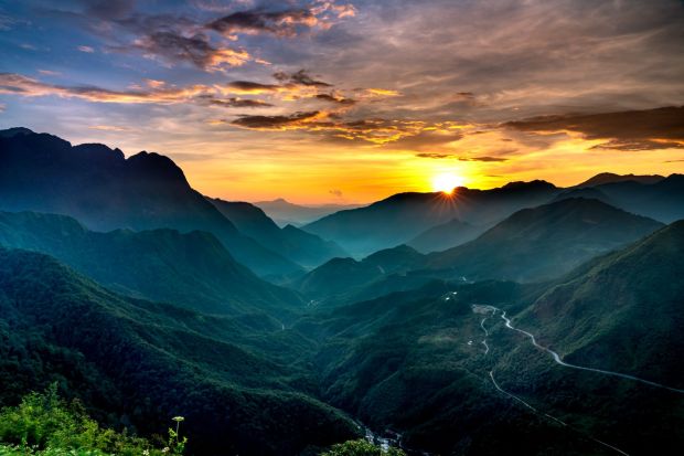 A breathtaking sunset over a mountainous landscape, with layered hills and valleys covered in lush greenery, as the sun dips below the horizon.